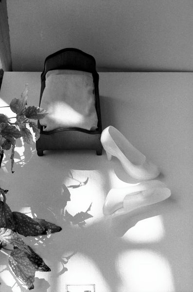 A pair of Barbie toy shoes is photographed in front of a miniature bed. Plants enter the frame from the left. The photograph is rendered in black and white.