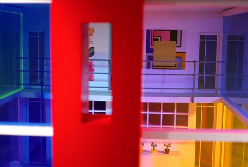 The coloured sliding windows of a modernist dollhouse offer a view into its interior. Past a red beam, the view opens onto a spacious foyer, bathed in purple light, where artworks are displayed.
