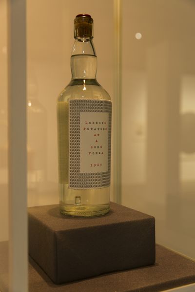 In a display case there is a bottle with a transparent liquid. The bottle label reads "LOBBING POTATOES AT A GONG VODKA 1969".