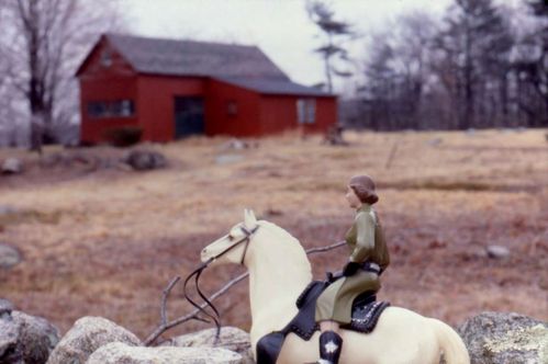 A female doll figure sits on the back of a toy horse. She is wearing a khaki-colored dress. Beyond a stone wall, a red barn with a black roof can be seen in soft focus.