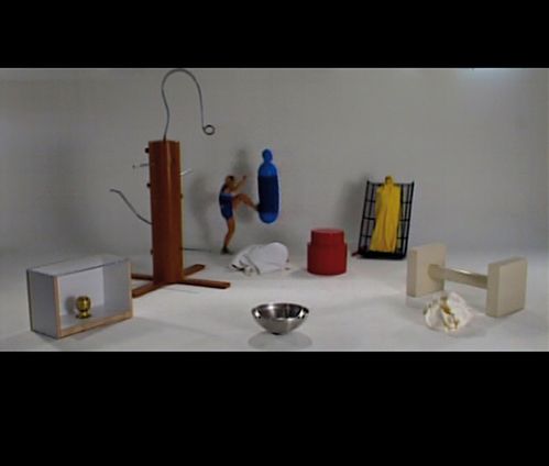 Video Screenshot: eine Art Bühne mit verschiedenen nicht klar identifizierenden Gegenständen. Inmitten eine Figur, die gegen eine Art Boxsack tritt