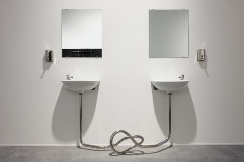 Two simple white sinks are mounted on a white wall. Plain mirrors hang above them. Stainless steel soap dispensers are installed to the left and right of the sinks. The drain pipes from the sinks do not go into the wall but instead connect in front of the sinks to form a knot.