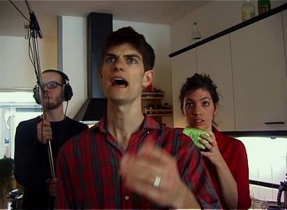 Filmstill. In einer Küche steht ein Mann mit kurzen dunklen Haaren und rot-blau kariertem Hemd in der Mitte des Bildes mit offenem Mund und gestikulierender Hand, als würde er jemandem was zurufen. Hinter ihm steht in der linken Bildhälfte ein bärtiger Mann mit Kopfhörern, der eine Mikrofonstange hält und auf der rechten Seite eine Frau mit roter Bluse, mit einem apfelgrünen rechteckigen Gegenstand in der Hand. Alle drei Personen blicken in die selbe Richtung, links am Betrachter vorbei.