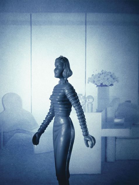Vor einem blau gehaltenen Hintergrund, der ein modern eingerichtetes Wohnzimmer andeutet, steht eine schmale Puppenfigur. Die Figur ist einfarbig gestaltet und im gleichen Blauton wie der Hintergrund glänzend lackiert. Sie trägt kurze Haare und einen Pullover mit Strickmuster.