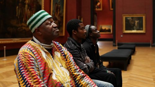 Three black men, one of them in colorful African clothing, sit next to each other on a museum bench and look in the same direction.