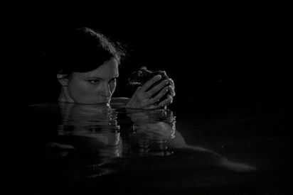 A woman is immersed up to her neck in a body of water in the dark and holds a rat with both hands in front of her face.