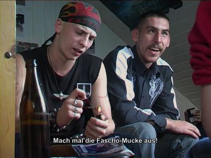 Zwei junge Männer sitzen nebeneinander. Einer ist tätowiert, hat ein KOptuch an und eine Kette mit einem silbernen Totenkopf-Anhänger. Er verbrennt etwas mit einem Feuerzeug. Der zweite träht ein zweifarbiges Hemd mit einem tribalen Muster und lacht. Ein Untertitel lautet: Mach mal die Fascho-Mucke aus!