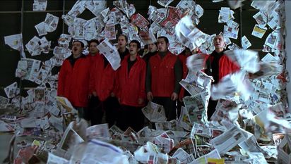A group of people in red jackets stand amidst sheets of newspaper whirling around in the air. It looks as if they are singing or shouting.