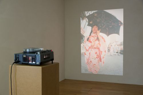 © YAYOI KUSAMA, photo: Thomas Dashuber A slide projector projects the image of an Asian woman in a pink kimono onto the wall. The woman is wearing an open umbrella covered with flowers.