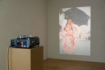 Diaprojektor projeziert das Bild einer asiatischen Frau im rosafarbenen Kimono an die Wand. Die Frau trägt einen offenen Schirm, der mit Blumen beklebt ist.