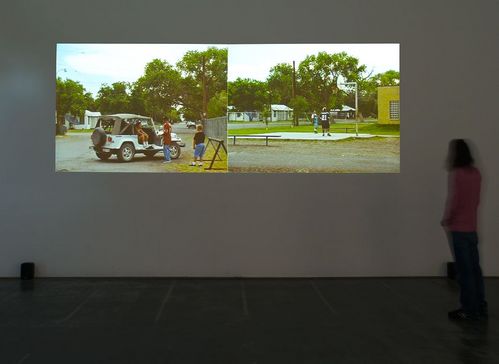 Two video projections are shown side by side on a wall in an exhibition space. They each show children and young people gathered around a white SUV and on a basketball court. 