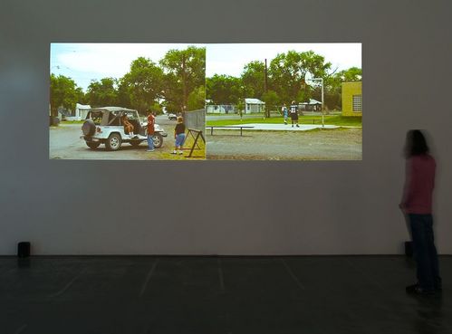 An einer Wand in einem Ausstellungsraum werden zwei Videoprojektionen nebeneinander gezeigt. Sie zeigen jeweils Kinder und Jugendliche um einen weißen Geländewagen versammelt und auf einem Basketballfeld. 