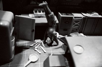 Die Schwarz-Weiß-Fotografie zeigt eine Puppenküche, leicht schräg von oben aufgenommen. Die Kühlschranktür steht offen, und auf dem Boden sind Geschirr und Besteck verstreut. Inmitten der Unordnung zwischen Küchenzeile und Küchentisch macht eine Puppenfigur einen Handstand.