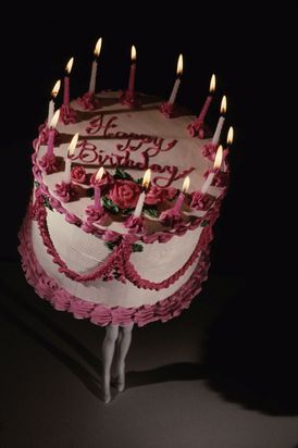 Eine pinke Geburtstagstorte  mit Kerzen und der Aufschrift „Happy Birthday“ läuft auf zwei Paar Beinen in hohen Schuhen. Die Aufnahme wurde aus erhöhter Perspektive fotografiert.