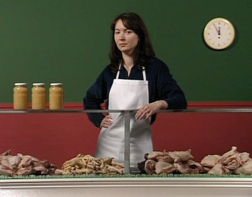 A woman wearing a white apron stands behind a counter with poultry. Behind her, the wall is painted horizontally green at the top and red at the bottom. There are three preserving jars with yellow contents on the counter