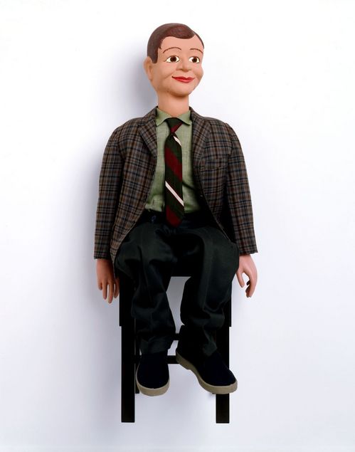 A ventriloquist dummy sits on  a chair mounted to the wall. It wears a plain, brown-toned checked jacket and a green shirt. Its shoes, like the trousers, are simple and dark, with a light contrasting sole. The dummy’s hair is neatly parted. Lines at the jaw indicate the movable mouth section.