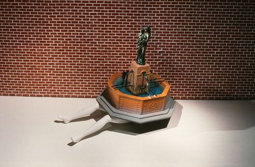 A toy fountain is mounted on lying doll legs. The fountain is octagonal. A pedestal extends from it, on which stands a black statue of a woman holding a child. The backdrop of this assemblage is a paper mock-up of a brick wall.