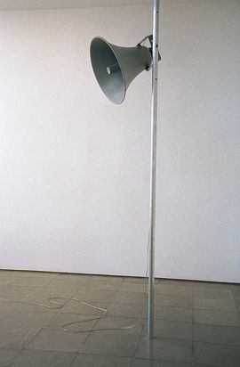 In einem Raum mit grauen Fließen ist vor einer weißen Wand ein Megaphon an einer silbernen Stange befestigt.