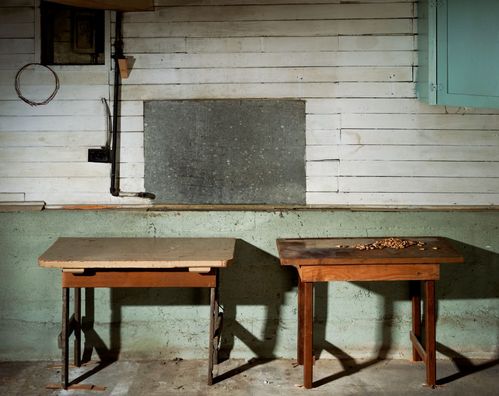 Zwei nebeneinanderstehende Holztische befinden sich vor einer Wand. Auf dem rechten Tisch liegen einige verstreute Bohnen. Der untere Teil der Wand wirkt wie Beton, während der obere Bereich aus weiß gestrichenem Holz besteht. Im oberen rechten Bildbereich hängt ein hellblaues Regal. Links oben ist eine Luke oder ein Fenster zu sehen, an dem ein rundes Seil befestigt ist. Hinter der Öffnung verläuft ein Rohr zu einem Betonabschnitt, der sich knapp oberhalb der Tische befindet.