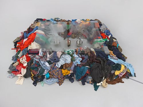 Eine quadratische Installation aus einem niedrigen Haufen bunter, zusammengewürfelter Kleidung, auf dem eine leicht beschlagene Glasplatte liegt; durch die Platte sind drei Wasserkessel sichtbar.