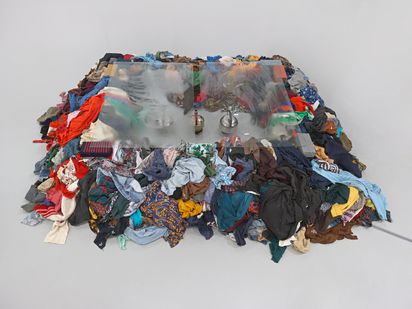 A large rectangular mound of mixed, colorful clothing piled on a white gallery floor with a frosted glass panel laid across the top and three metal kettles visible beneath the glass.