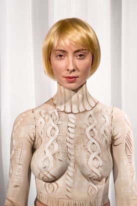 Vor einem weißen Hintergrund wurde das Porträt einer Frau mit blondem Bob fotografiert. Sie trägt scheinbar einen Kabelstrickpullover. Bei näherem Hinsehen wird jedoch deutlich, dass dieser lediglich auf ihren nackten Oberkörper aufgemalt ist.