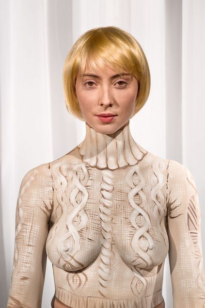 A portrait of a woman with a blonde bob is photographed against a white background. She appears to be wearing a cable-knit sweater. On closer inspection, however, it becomes clear that the garment is merely painted onto her bare upper body.