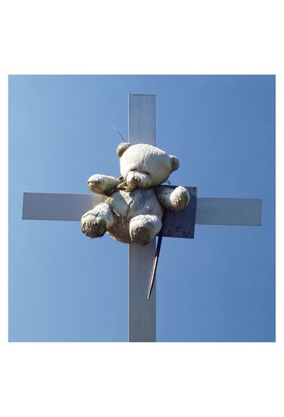 Photographs of white grave crosses against blue cloudless sky. All have simple memorial plates made of sheet metal, on which the names and life dates of the deceased are handwritten. Children's toys or stuffed animals are attached to many of the crosses. Both the crosses and the toys have been soiled by the weather and have come down. In some photos, wild grasses and yellow flowers protrude into the picture from below.