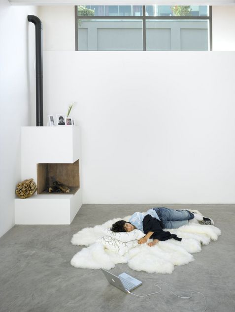 Ein junger Mann liegt ausgestreckt auf einem Lammfellteppich. Er trägt Jeans, ein Polohemd und Converse-Schuhe, die er teilweise ausgezogen hat. Vor ihm steht ein Laptop, den er nicht ansieht. Im Hintergrund befindet sich ein ungenutzter Kamin mit persönlichen Fotografien in weißen Rahmen und einer Vase mit Blumen auf dem Sims.