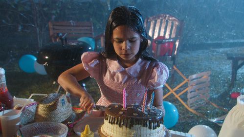 In the pouring rain, a girl with dark hair and a pink dress cuts a birthday cake