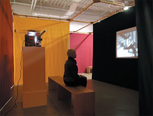 A visitor sits in a room separated by colorful fabric panels and watches a video projection in black and white