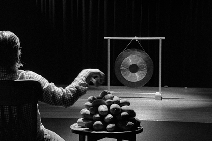 Black and white shot. In the left corner sits a man with shoulder-length hair with the back to the viewer on a wooden chair. Next to him is a small round high table, on which neatly stacked potatoes lie. The man reaches with his right hand to the top of the pile. In front of him is a metal gong in front of a dark curtain.