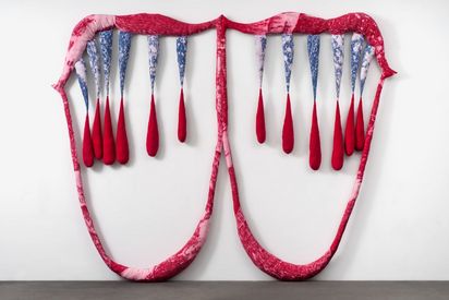  Two connected, mouth-like forms are made of pink, padded fabric strips. From the upper form, several teardrop-shaped, filled fabric elements hang down, their tips tinted red.
