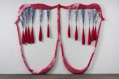 Two connected, mouth-like forms are made of pink, padded fabric strips. From the upper form, several teardrop-shaped, filled fabric elements hang down, their tips tinted red.