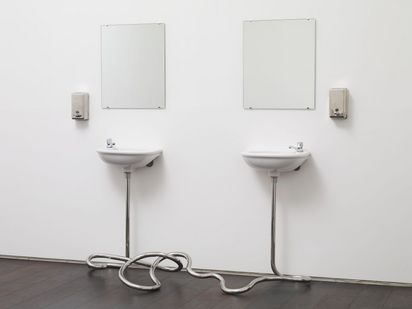 Zwei einfache weiße Waschbecken sind an einer weißen Wand montiert. Darüber hängen schlichte Spiegel. Links und rechts der Waschbecken sind Edelstahl-Seifenspender angebracht. Die Abflussrohre der Waschbecken führen nicht in die Wand, sondern verbinden sich vor den Becken zu einem Knoten. 