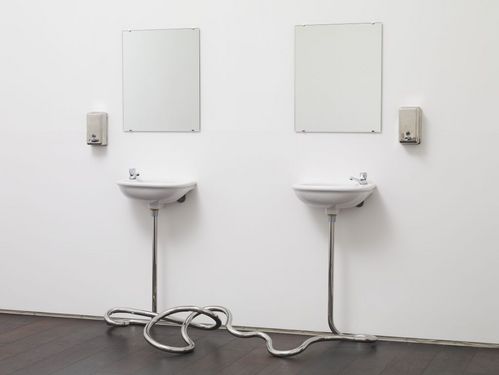 Zwei einfache weiße Waschbecken sind an einer weißen Wand montiert. Darüber hängen schlichte Spiegel. Links und rechts der Waschbecken sind Edelstahl-Seifenspender angebracht. Die Abflussrohre der Waschbecken führen nicht in die Wand, sondern verbinden sich vor den Becken zu einem Knoten. 