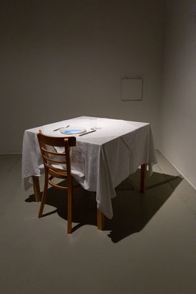 Ein Stuhl steht vor einem viereckigen Tisch mit weißem Tischtuch. Auf dem Tisch steht ein Teller, in den ein Bildschirm eingebaut ist.