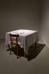 Ein Stuhl steht vor einem viereckigen Tisch mit weißem Tischtuch. Auf dem Tisch steht ein Teller, in den ein Bildschirm eingebaut ist.