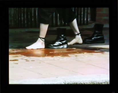 Nackte Füßen schreiten über ein Trottoir. An den Fesseln sind mit Schnürsenkeln schwarze Dr. Martens Boots befestigt, die sie Schritt für Schritt hinter sich herziehen.