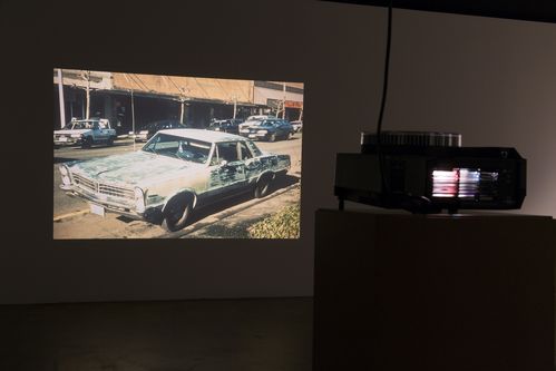 In einem dunklen Raum projiziert ein Dia-Projektor das Bild eines auf einer Straße geparkten altmodischen Automodells an eine Wand.
