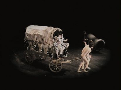 Standbild aus einem Video. Gezeigt wird eine Inszenierung auf einer Bühne: Zwei Personen mit hautfarbenen Kostümen, die Nacktheit symbolisieren, ziehen einen Planwagen. Auf diesem Sitzen zwei Personen, im Hintergrund ist eine Kanone zu sehen.