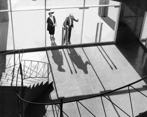 Black and white shot of a man and a woman standing in front of a glass door, as if trying to gain access to the room behind it.
