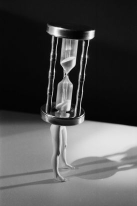Auf Puppenbeinen ist eine Sanduhr montiert, die den Oberkörper der Figur bildet. Die Fotografie ist in Schwarz-Weiß gehalten und zeichnet sich durch einen schwarzen Hintergrund sowie starke Schatten aus.