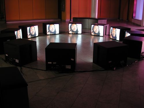 Many monitors form a circle on the floor. They all show the same image of a bearded man
