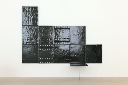 Schwarze, glänzende Metallplatten mit Nieten und unterschiedlichen Oberflächenstrukturen, die zu einer geometrischen Wandinstallation zusammengesetzt sind. Unten rechts ragt eine einzelne Platte im rechten Winkel ab, sie wird von einem dünnen Metallstab getragen.
