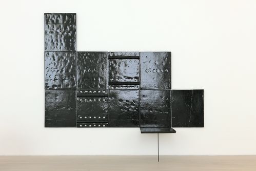 Schwarze, glänzende Metallplatten mit Nieten und unterschiedlichen Oberflächenstrukturen, die zu einer geometrischen Wandinstallation zusammengesetzt sind. Unten rechts ragt eine einzelne Platte im rechten Winkel ab, sie wird von einem dünnen Metallstab getragen.