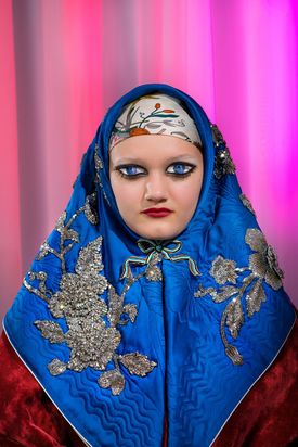 Vor einem pinken Hintergrund wurde das Porträt einer Frau in traditioneller Kleidung fotografiert. Ein leuchtend blaues, mit Silberapplikationen geschmücktes Tuch rahmt ihren Kopf. Unter dem Kinn ist es mit einer kleinen Schleife gebunden. Die Frau hat die Augen geschlossen, doch auf ihre geschlossenen Augenlider wurden mit Schminke Augen aufgemalt. Diese blicken den Betrachter mit stechendem Ausdruck an.