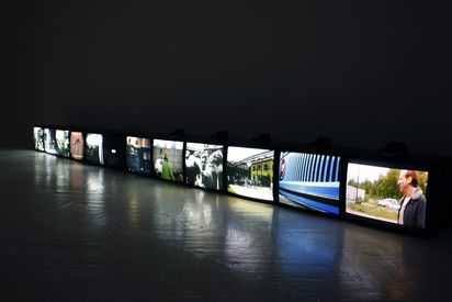 12 different TV screens lie side by side on the floor. They each show different film scenes, some of which feature people.