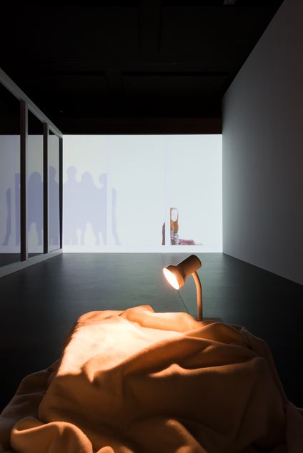 Room installation with video projection on the background. It shows a kneeling girl on the floor, with the silhouettes of several standing people depicted as shadows next to her. In the foreground, a blanket lies on the floor, illuminated by a bedside lamp.