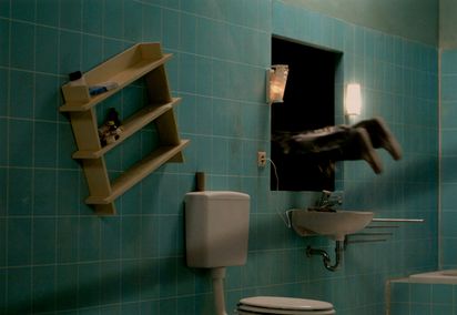 In a bathroom, instead of the mirror above the washbasin, there is a hole with a pair of human feet sticking out of it, as if a person had jumped in. Next to it, a shelf hangs askew.